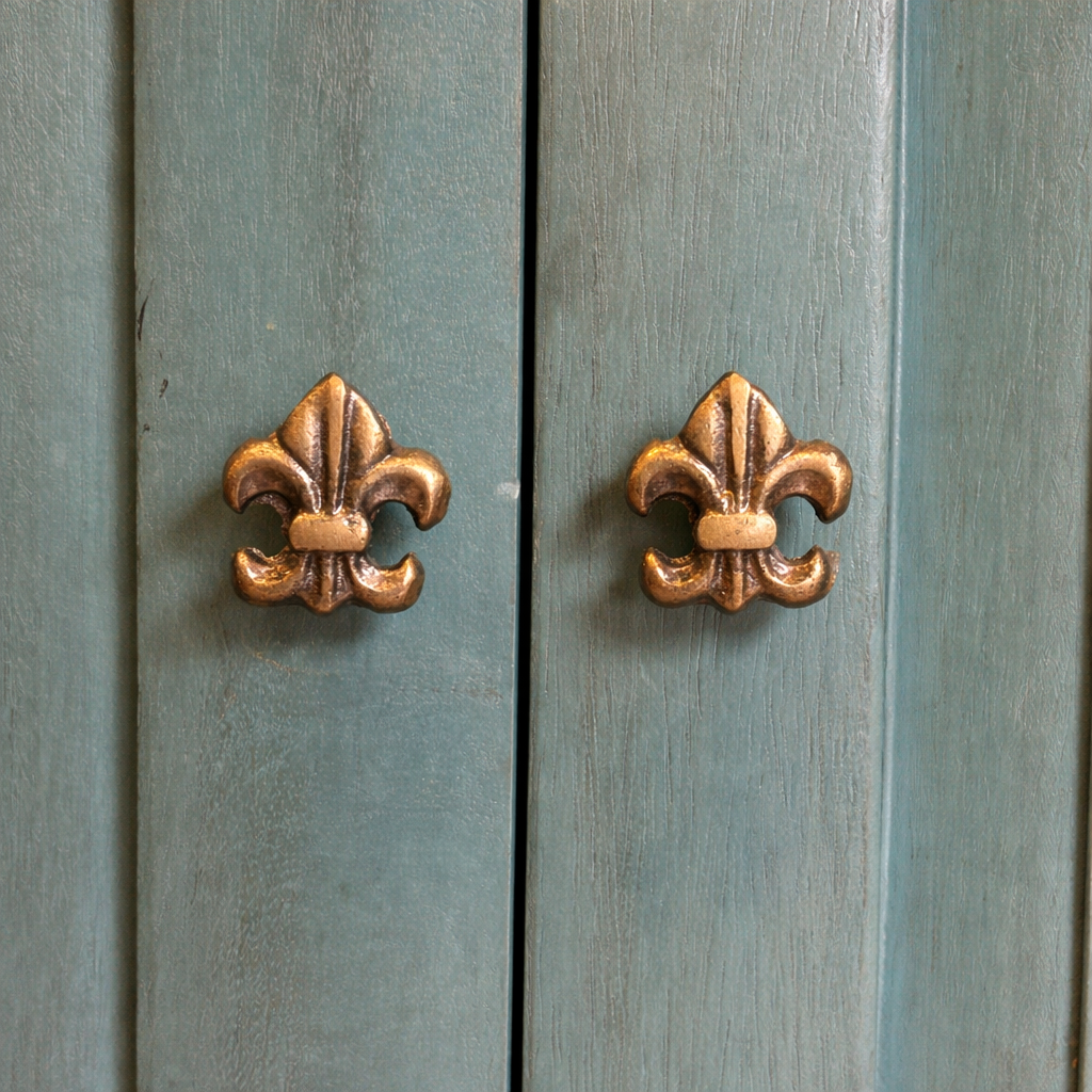 Close-up of antique brass fleur-de-lis cabinet knobs on a distressed turquoise wooden cabinet door, highlighting textured finish and vintage detailing.