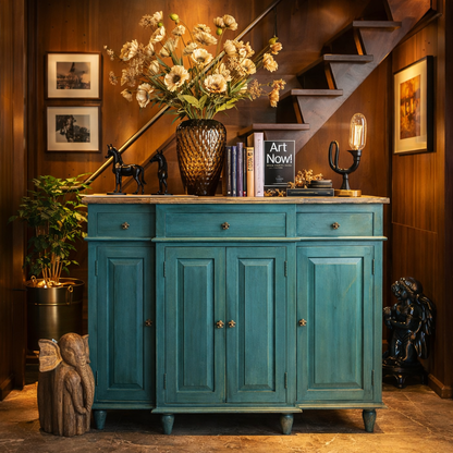 Distressed teal wooden sideboard with antique brass knobs styled in a luxury entryway with amber glass vase of flowers, decorative books, sculptural accents, and warm wood-paneled background.