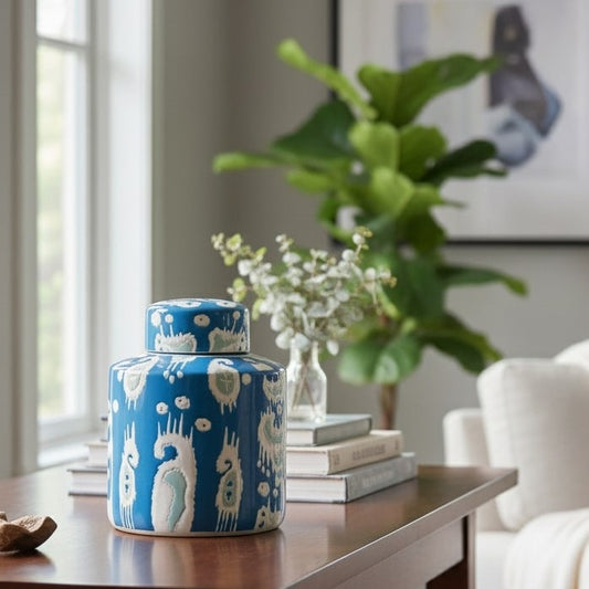 A premium handpainted blue ceramic ginger jar with a white ikat pattern, sitting on a stack of books on a wooden console table, styled with greenery and a fiddle leaf fig in a bright, elegant living room.
