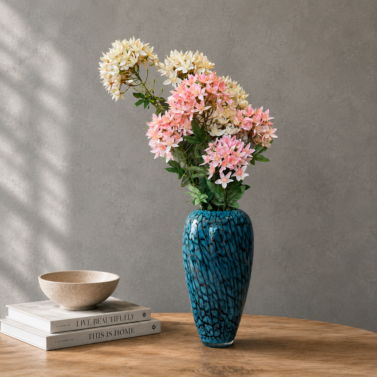 Blue textured glass vase with pink and cream artificial flowers styled on a wooden table against a grey wall in a modern minimalist interior.