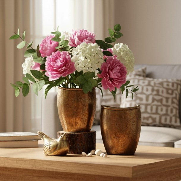 Two textured bronze vases on a light wood table; one holds a large bouquet of peonies and hydrangeas, accompanied by a small gold bird figurine and white pebbles.