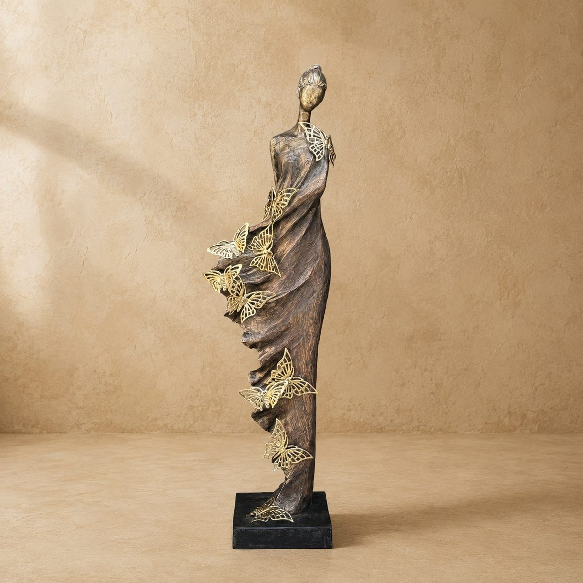 Butterfly Lady decorative sculpture with intricate gold butterflies and an antique bronze finish displayed against a warm beige Venetian plaster wall in soft natural daylight.