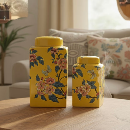 Two yellow rectangular ceramic jars with floral patterns displayed on a wooden coffee table in a sunlit, cozy living room setting.
