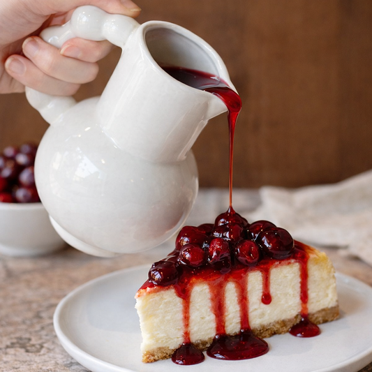A white ceramic creamer pouring vibrant red cherry sauce over a slice of New York-style cheesecake.
