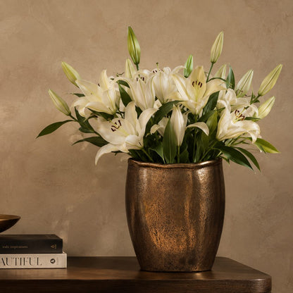 Antique bronze handcrafted decorative metal vase styled with elegant white lilies on a wooden console table.