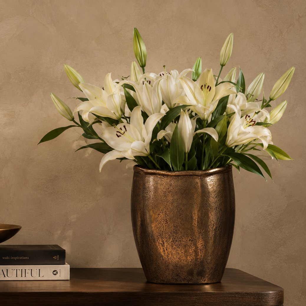 Antique bronze handcrafted decorative metal vase styled with elegant white lilies on a wooden console table.
