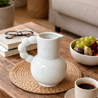 A glossy white ceramic pitcher with a unique bubble-style sculptural handle sitting on a woven coaster on a wooden coffee table.