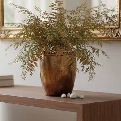 A delicate green fern plant arranged in a textured bronze vase, styled on a wooden table with a gold mirror blurred in the background.
