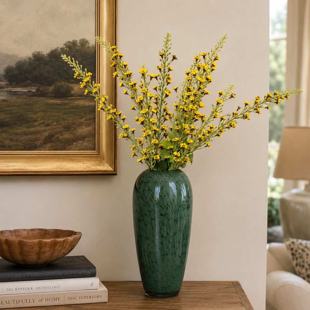 Elegant green glass vase with delicate yellow artificial flowers styled on a wooden console table in a warm contemporary living room setting.
