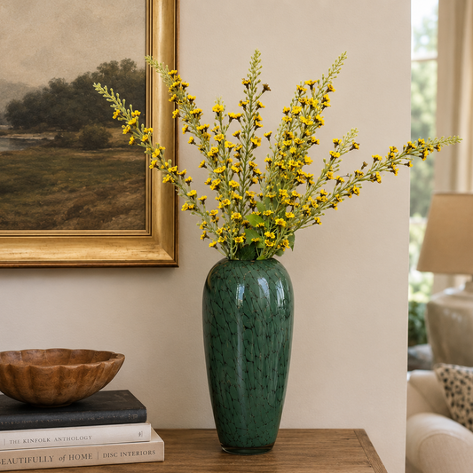 Elegant green glass vase with delicate yellow artificial flowers styled on a wooden console table in a warm contemporary living room setting.