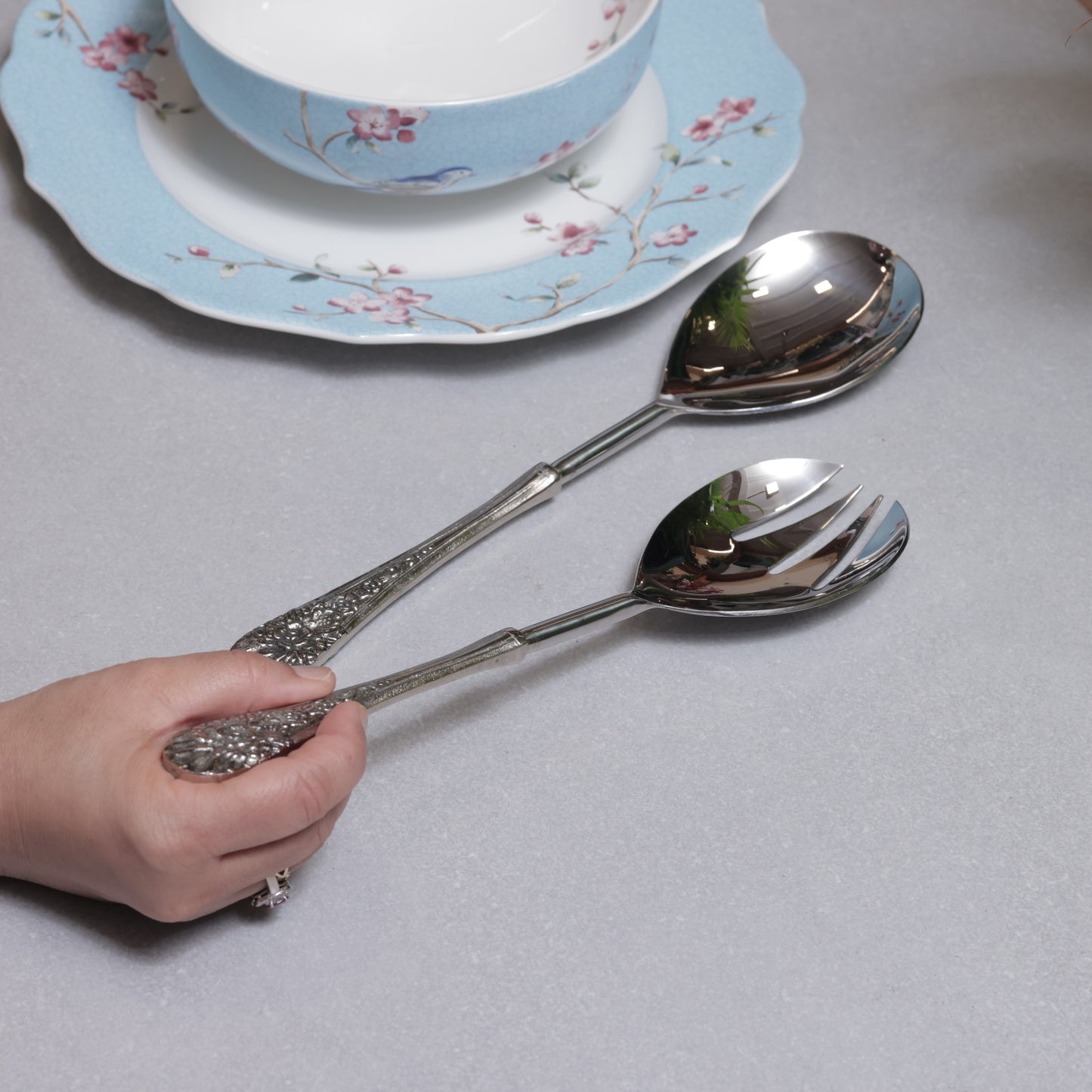 A person’s hand holding a pair of ornate silver-plated salad servers over a light grey surface next to a blue floral ceramic bowl.