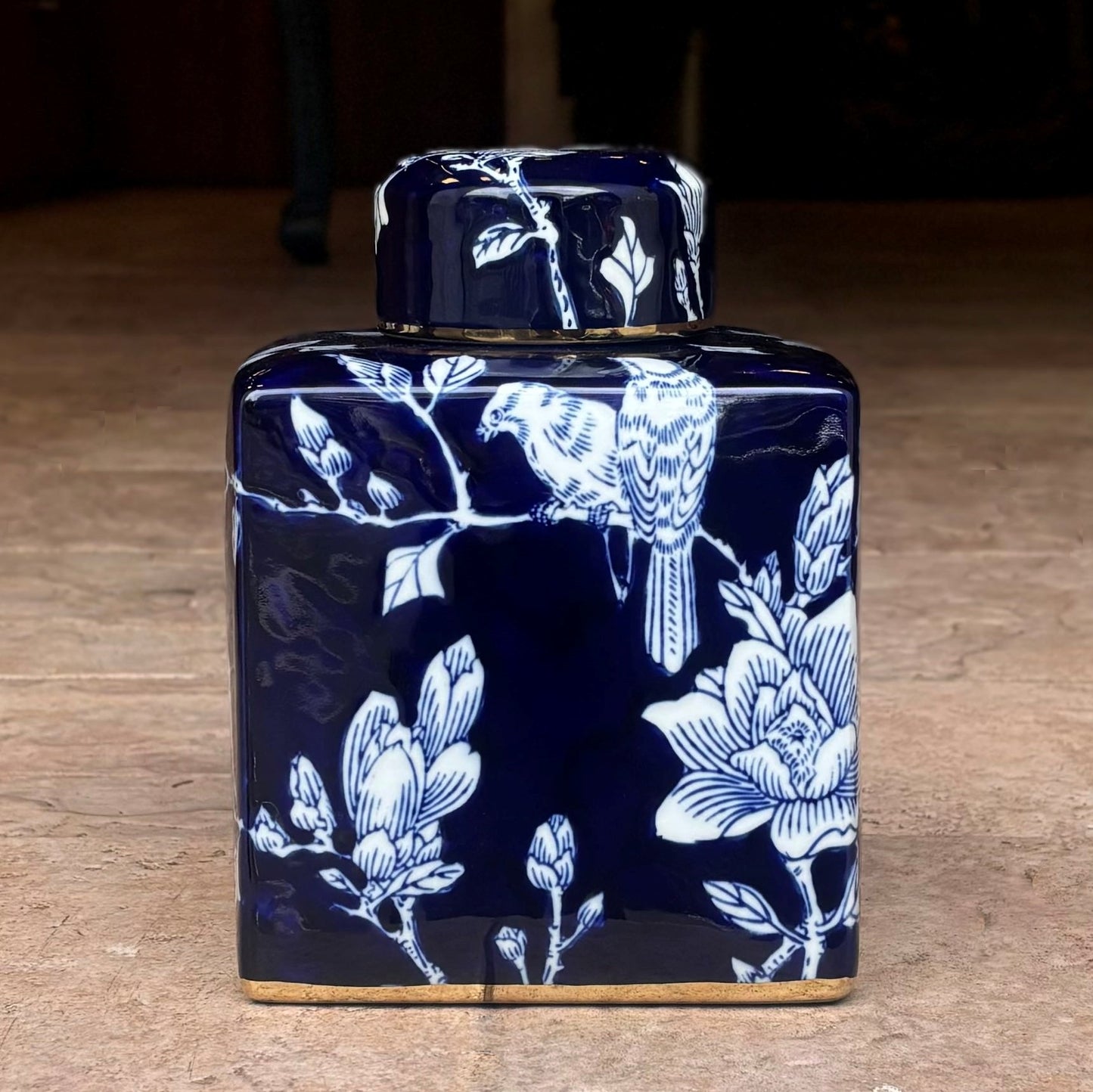 Close-up of a square cobalt blue porcelain decorative jar featuring a hand-painted white bird perched on a magnolia branch, accented with a luxurious gold leaf finish at the base.