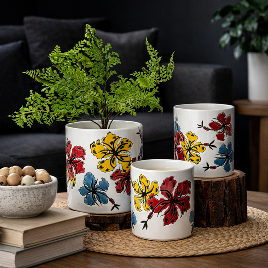 Set of three hand-painted ceramic floral planters with green plant on a wooden coffee table in a living room with dark sofa and wall in natural light.