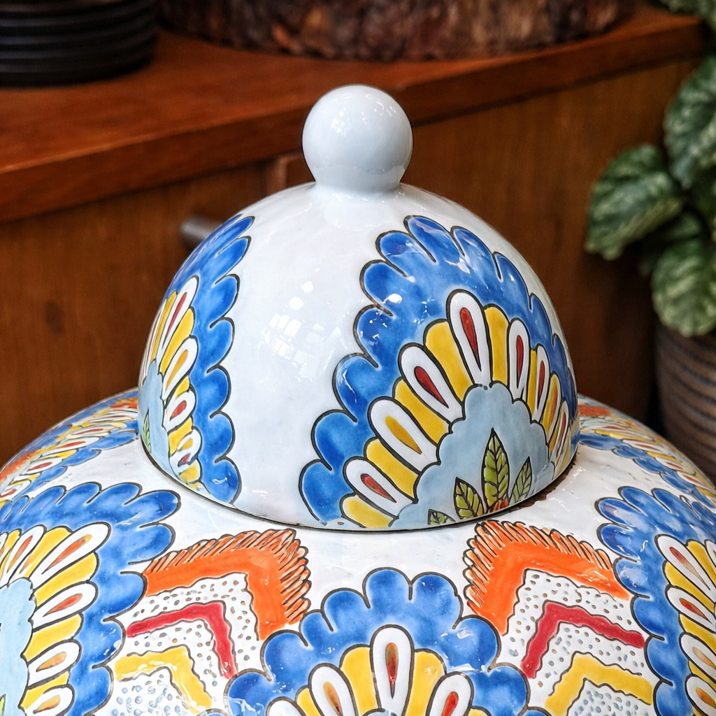 Close-up lifestyle image of a hand-painted ceramic ginger jar lid featuring intricate blue, yellow, and orange floral detailing with a glossy white finial in a warm wooden interior setting.