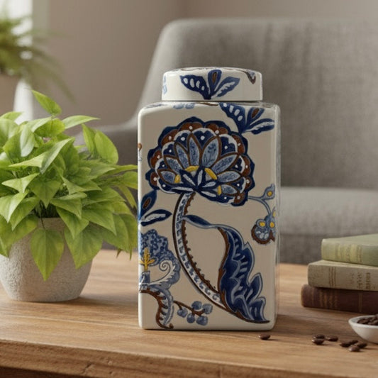 A rectangular hand-painted ikat decorative ceramic jar with blue and brown floral patterns, styled on a wooden table next to a green potted plant and vintage books in a bright, modern living room.