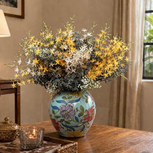 Close-up lifestyle image of a handpainted floral ceramic vase with yellow and white artificial blooms arranged on a wooden tabletop in a warm, elegant interior setting.