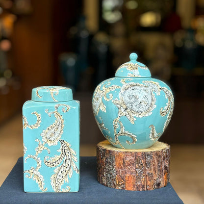 A round, bulbous ceramic ginger jar with a matching lid, displayed on a natural wood log slice, featuring a detailed paisley design on a turquoise background.