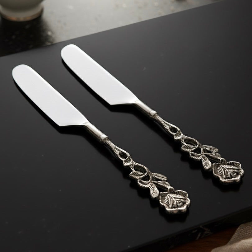 A pair of handcrafted silver butter knives with floral rose-patterned handles resting on a sleek black marble slab.