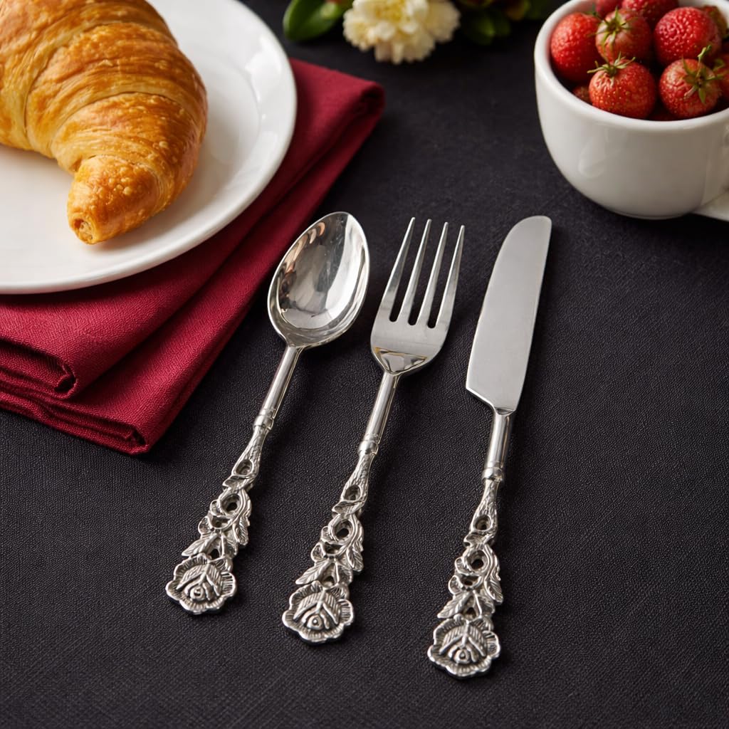 A complete artisanal flatware set including a spoon, fork, and knife with floral etched handles, styled on a dark linen surface with a fresh croissant and strawberries.