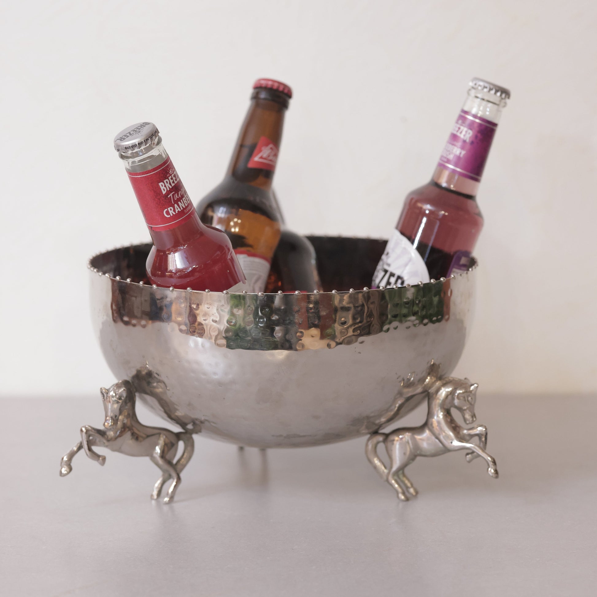 A hammered silver metal bowl used as an ice bucket for drinks, supported by three detailed rearing horse figurines on a neutral surface.