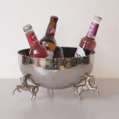 A hammered silver metal bowl used as an ice bucket for drinks, supported by three detailed rearing horse figurines on a neutral surface.