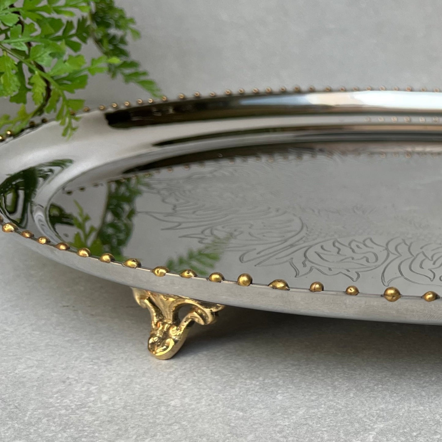 Close-up of a high-shine silver-plated oval tray featuring hand-etched floral patterns, a decorative gold beaded edge, and ornate brass-finished feet on a neutral textured surface.