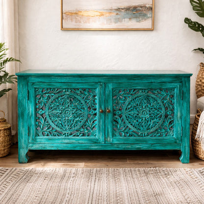 Square lifestyle image of a distressed turquoise wooden cabinet with intricate hand-carved mandala panels and brass knobs, styled against a neutral wall with full product visibility.