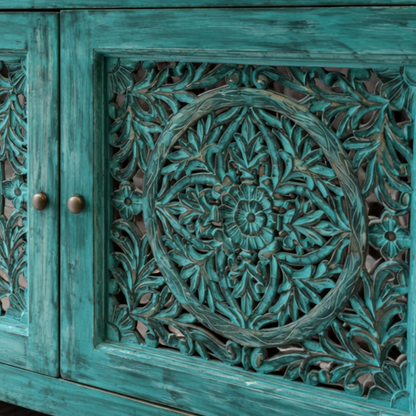Close-up detailing of intricate hand-carved floral cut work on a distressed turquoise wooden cabinet door, highlighting fine craftsmanship and textured finish.