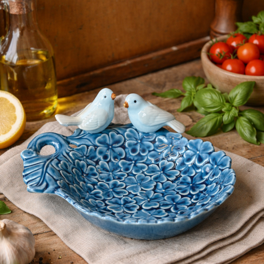 Hand-painted blue ceramic decorative bowl with twin bird figurines and embossed floral design, styled on a rustic kitchen table with basil, tomatoes, olive oil and lemon, artisan home décor accent.
