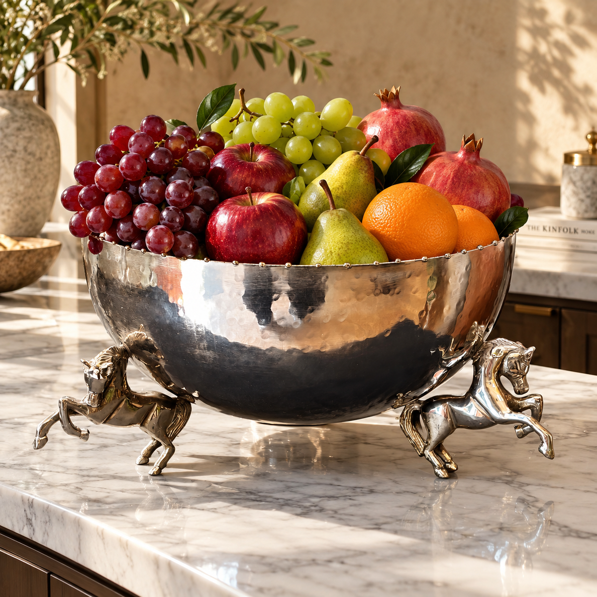 Luxury silver decorative horse leg bowl filled with fresh apples, grapes, pears, oranges and pomegranates on a marble countertop.