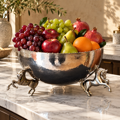 Luxury silver decorative horse leg bowl filled with fresh apples, grapes, pears, oranges and pomegranates on a marble countertop.