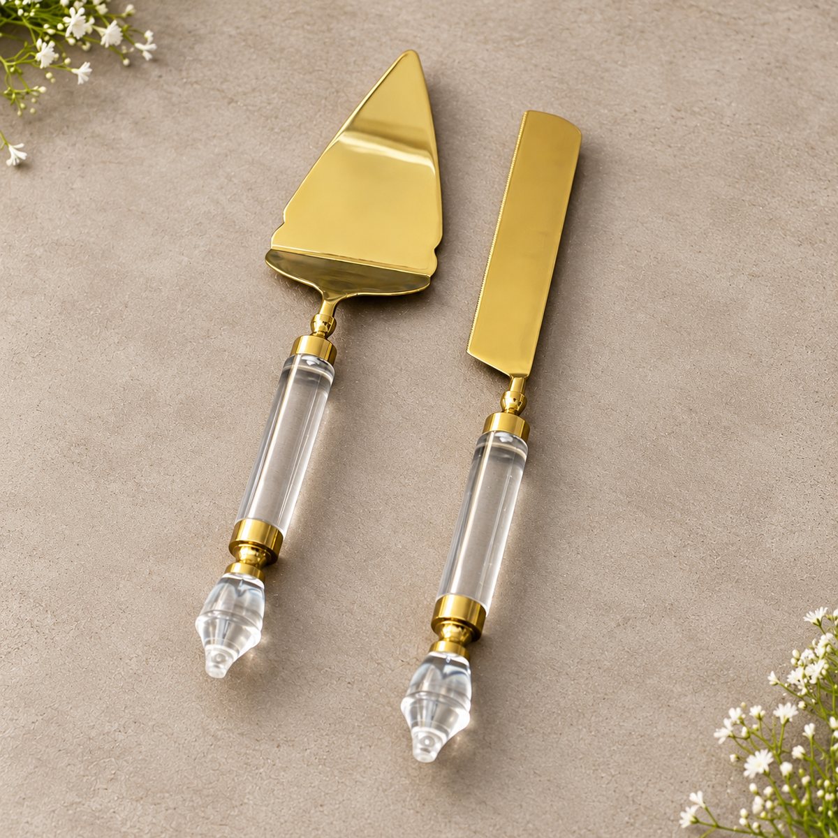 Luxury gold cake server and serrated knife set with crystal-style clear acrylic handles resting on a neutral stone backdrop with soft shadows.