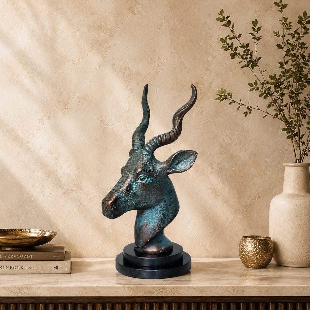 Luxury antique bronze antelope head sculpture displayed on a premium fluted console against a warm beige Venetian plaster wall with natural light, styled with designer books, a ceramic vase, and elegant home décor accessories.
