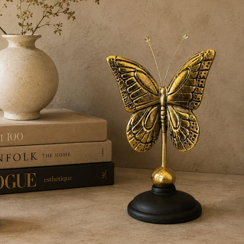 Antique brass butterfly sculpture on a black base displayed beside stacked coffee table books, a ceramic vase, and a decorative candle, showcasing its size in a luxury home décor setting.