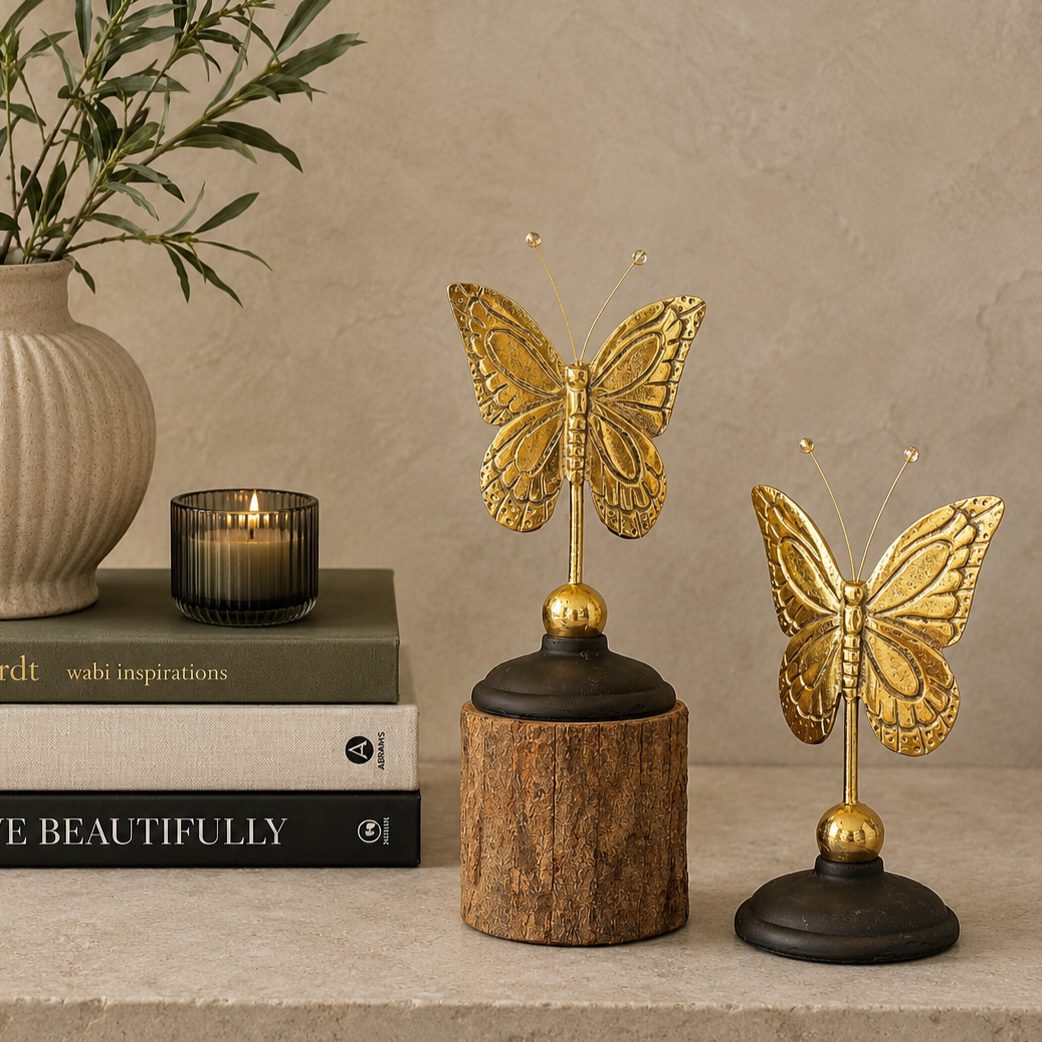 Antique brass butterfly figurines displayed beside stacked luxury coffee table books, a ceramic vase, and a decorative candle on a warm beige backdrop, showcasing their size and elegant home décor styling.