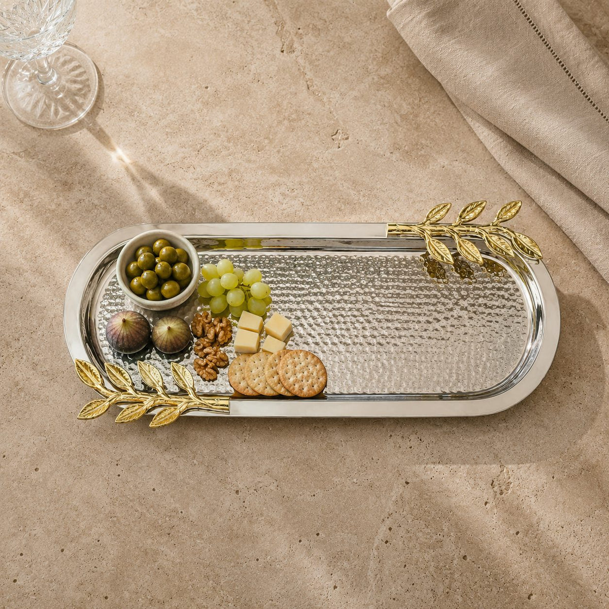 Leaf Handle Hammered Serving Tray with gold leaf handles styled as a luxury charcuterie platter featuring cheese, grapes, olives, figs, walnuts, and crackers on an elegant stone tabletop.