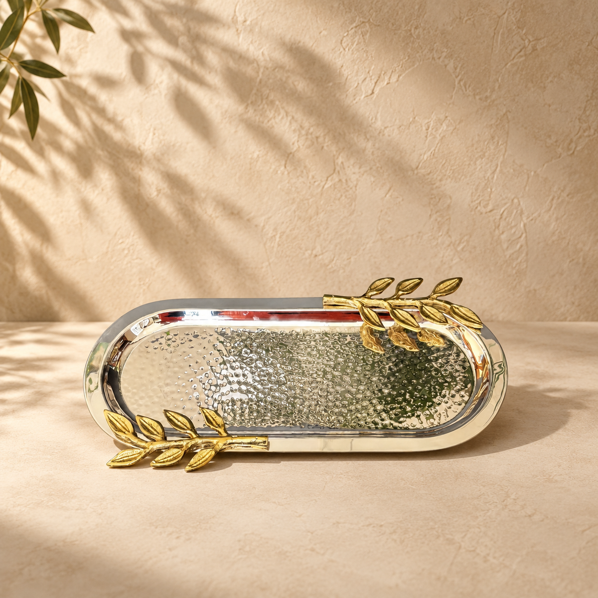 Premium hammered stainless steel serving tray with gold leaf handles displayed on a luxury beige studio background.