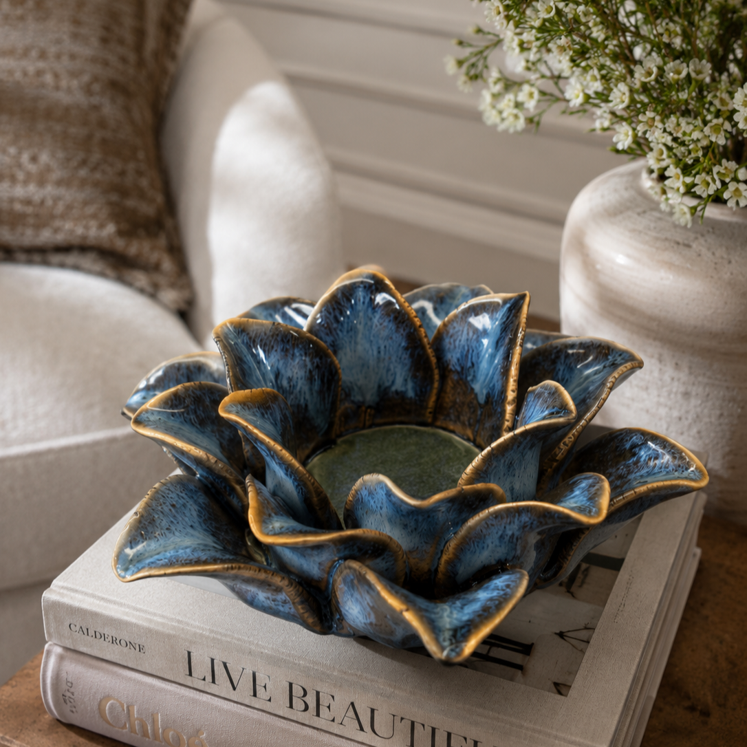 Close-up lifestyle image of a handcrafted blue ceramic flower-shaped tealight holder placed on stacked books beside floral décor in a cozy modern living space.