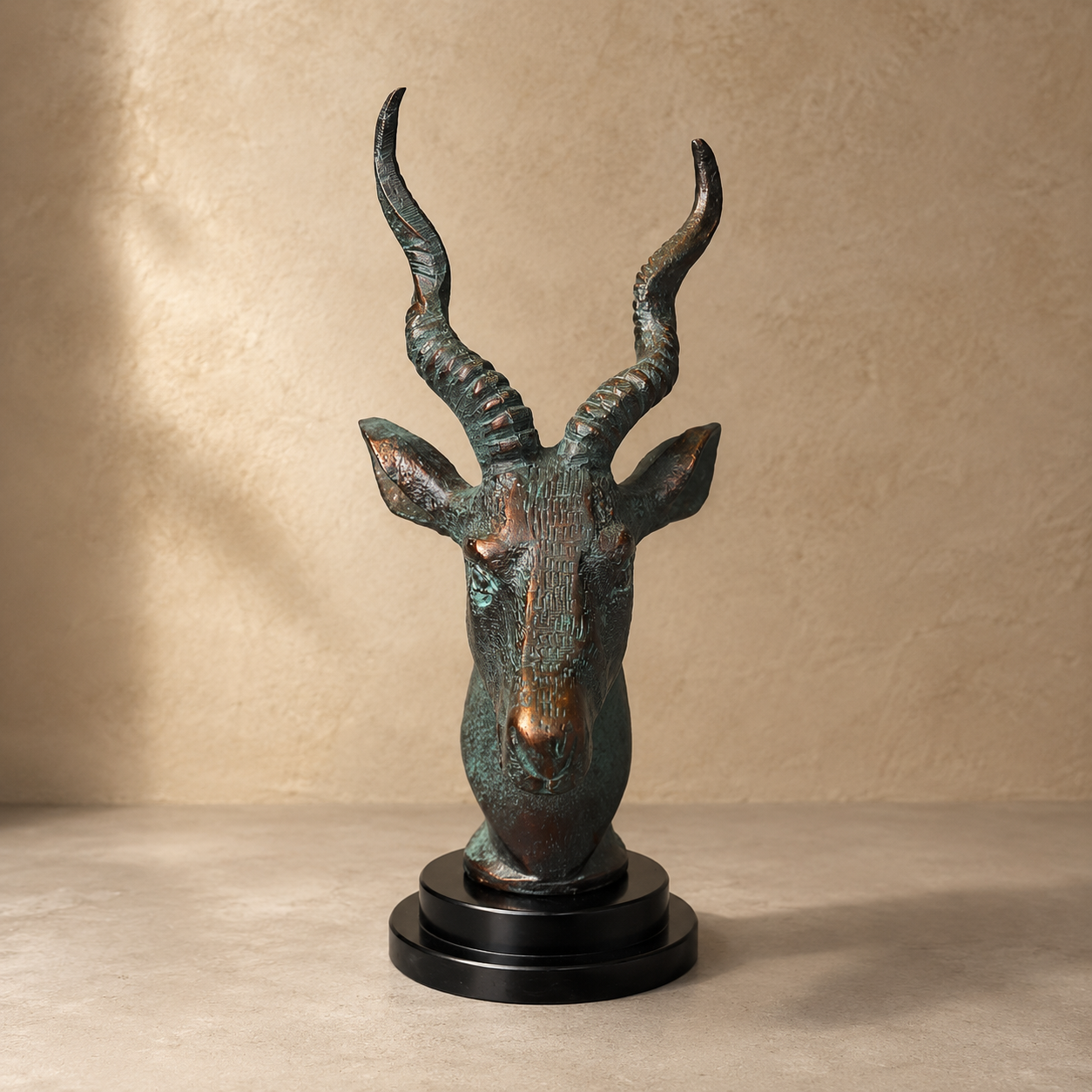 Luxury patina bronze antelope head sculpture on a black pedestal, photographed from the front against a warm beige Venetian plaster backdrop in a premium studio setting.