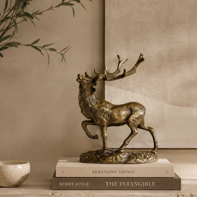 Luxury bronze stag decorative sculpture styled on stacked coffee table books atop a travertine console, with olive branches and a warm beige textured wall in a premium contemporary home interior.