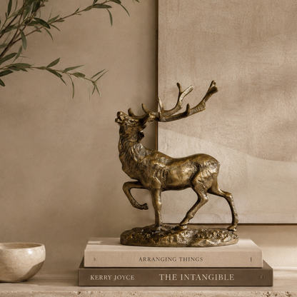 Luxury bronze stag decorative sculpture styled on stacked coffee table books atop a travertine console, with olive branches and a warm beige textured wall in a premium contemporary home interior.