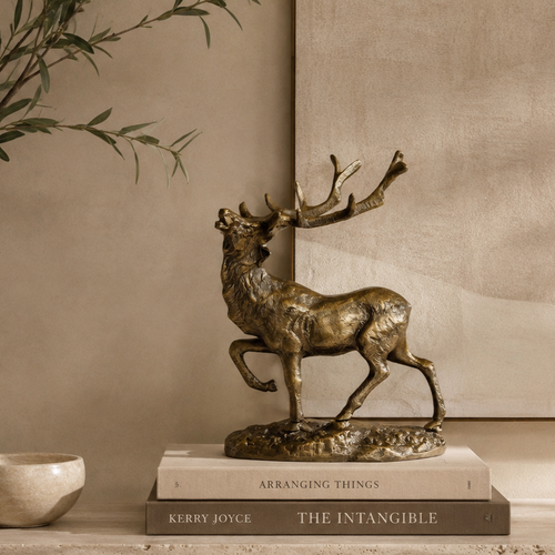 Luxury bronze stag decorative sculpture styled on stacked coffee table books atop a travertine console, with olive branches and a warm beige textured wall in a premium contemporary home interior.