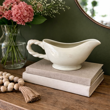 Cream ceramic gravy boat styled as a decorative accent on stacked neutral books, placed on a wooden console table against a dark olive green wall with blush pink flowers in the background.