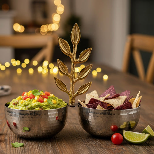 Stylish hammered metal double snack bowl with gold leaf handle serving guacamole and colorful tortilla chips on a wooden dining table.