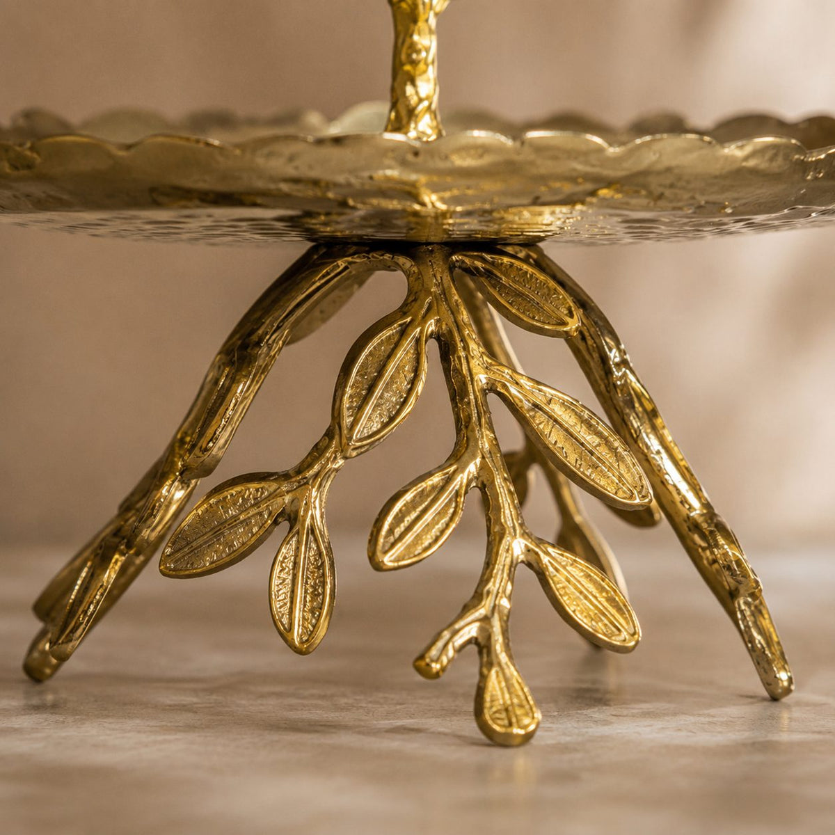 Close-up of the handcrafted gold leaf base of a luxury two-tier serving stand with intricate botanical branch detailing and hammered metal finish.