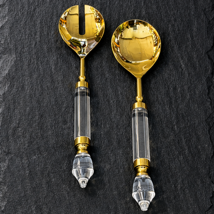 Premium gold salad server set with mirror-polished stainless steel heads and clear acrylic handles, displayed on a dark black slate background
