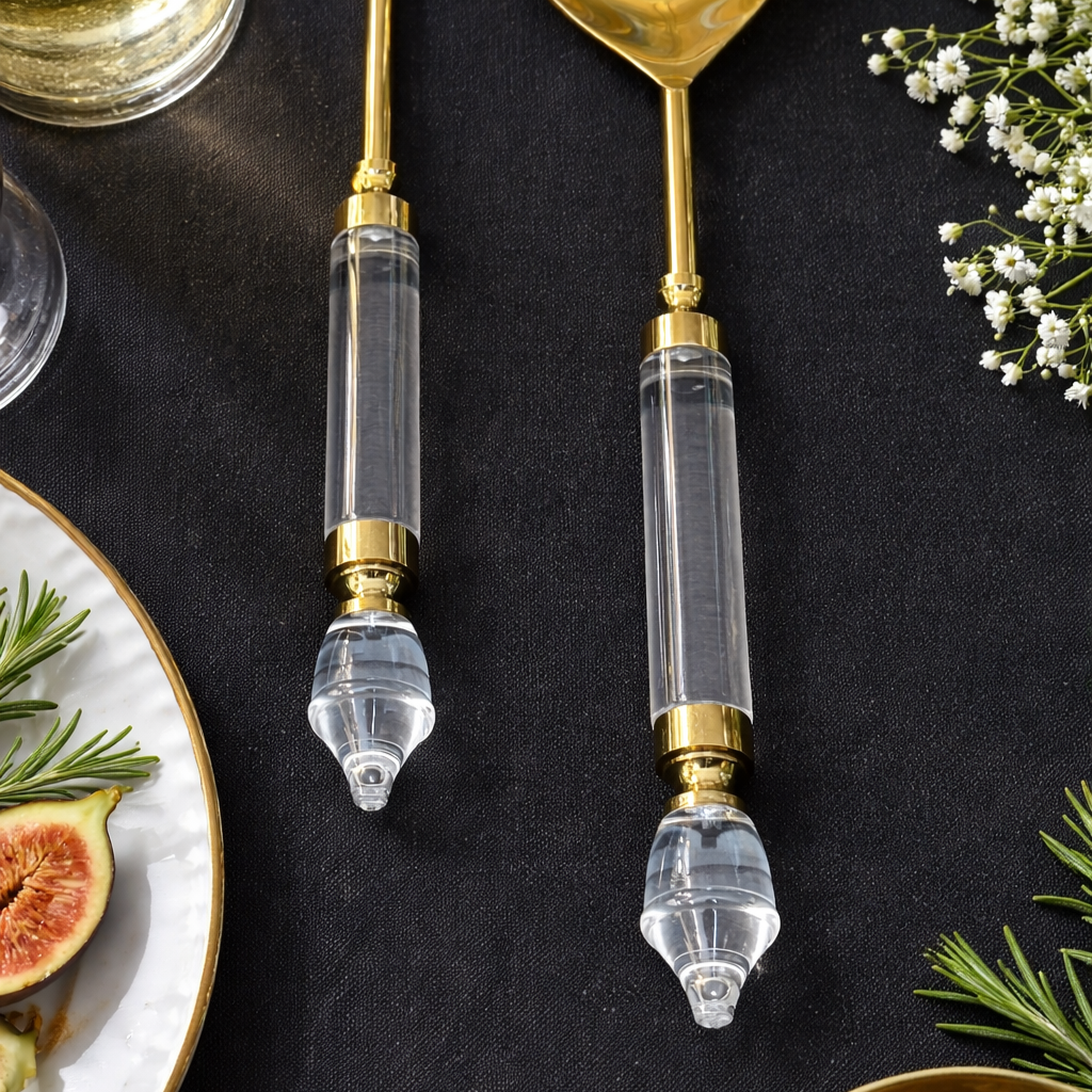 Close-up of premium gold salad serving spoon and fork with clear acrylic handles styled on a dark luxury dining table setting with figs, rosemary, and wine
