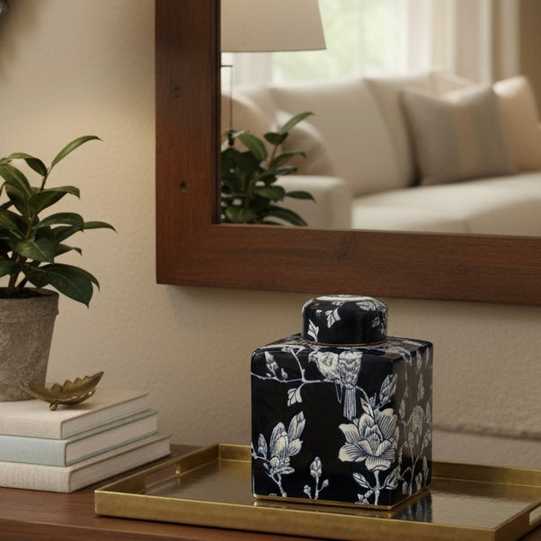 A premium blue and white Chinoiserie jar styled on a brushed gold tray next to a stack of books and a potted plant, reflected in a large wooden-framed mirror in a bright living room.