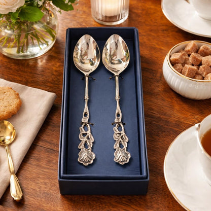 Two silver rose-handled dessert spoons in a navy blue gift box on a wooden table set for tea with cookies and sugar cubes.
