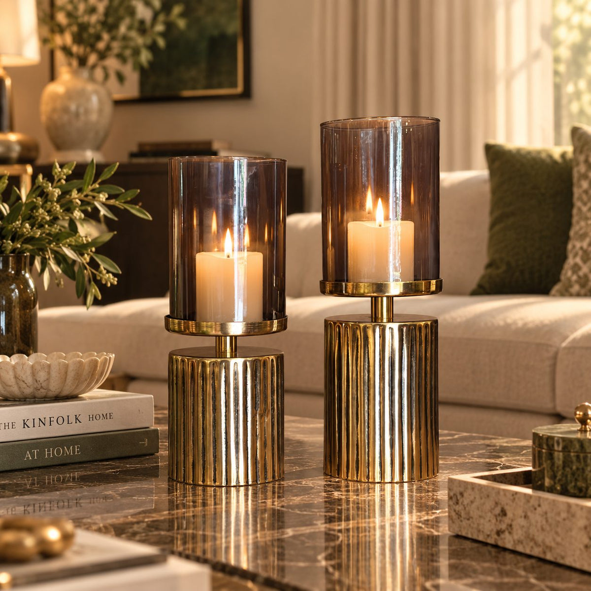 Luxury smoked glass hurricane candle holder set with lit pillar candles styled on a marble coffee table in an elegant living room interior.